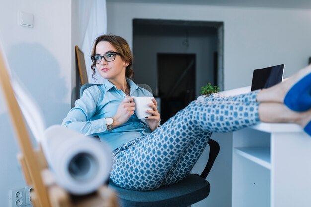 Durchdachte junge Geschäftsfrau, die Kaffeetasse sich entspannt auf einem Bürostuhl hält