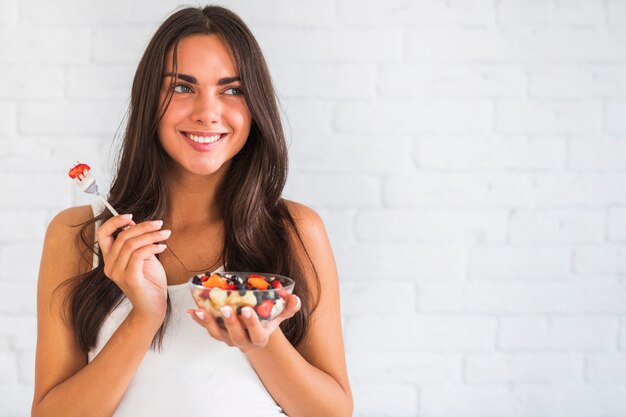 Durchdachte lächelnde junge Frau, die Schüssel Fruchtsalat hält