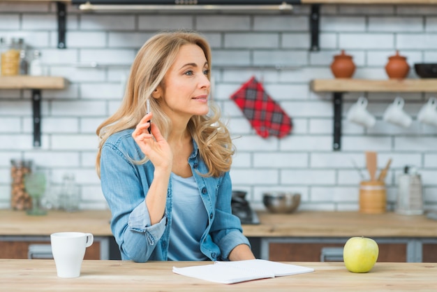 Durchdachter blonder Holdingbehälter der jungen Frau in der Hand mit Buch; Apfel- und Weißschale auf Holztisch