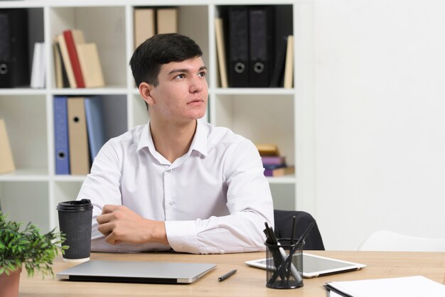 Durchdachter junger Geschäftsmann, der am Schreibtisch weg schaut sitzt