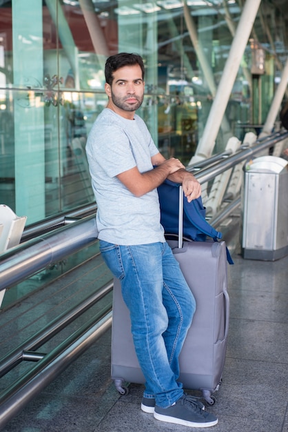 Durchdachter junger Mann, der auf Metallgeländer am Flughafen sich lehnt