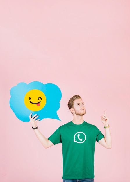 Durchdachter mann, der aufwärts beim anhalten des blinzelns der emoji spracheblase zeigt