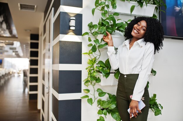 Durchscheinende afroamerikanische Geschäftsfrau mit Afro-Haaren, weißer Bluse und grünen Hosen, posiert im Café mit Handy zur Hand