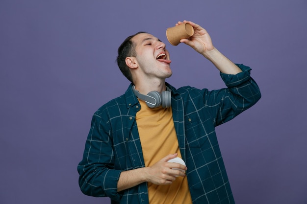 Durstiger junger männlicher Student, der Kopfhörer um den Hals trägt und eine Kaffeetasse aus Papier über dem Mund hält, und seine Kappe in einer anderen Hand, die in die Tasse mit offenem Mund schaut und die Zunge isoliert auf violettem Hintergrund zeigt