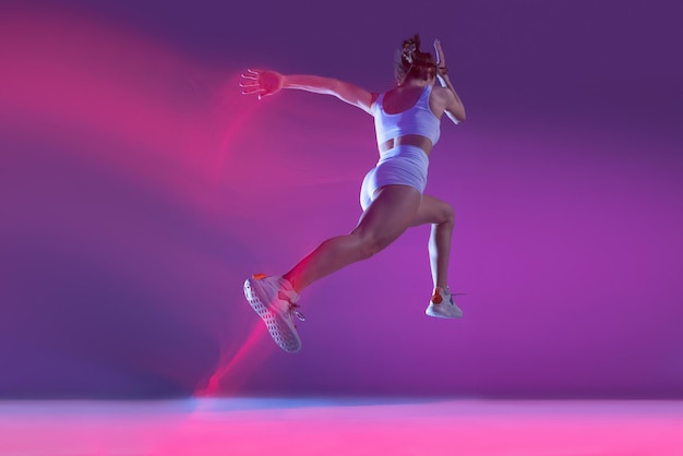 Dynamisches Porträt einer jungen sportlichen Frau, die isoliert über violettem Hintergrund in Neon mit gemischten Lichtern läuft