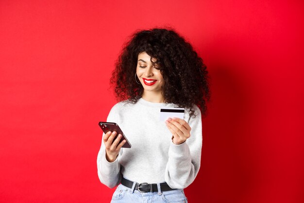 E-Commerce- und Online-Shopping-Konzept. Attraktive kaukasische Frau, die für den Kauf im Internet bezahlt, Smartphone und Kreditkarte hält, roter Hintergrund