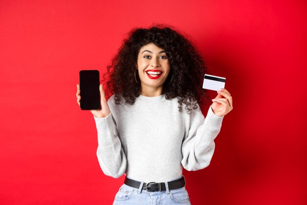 E-Commerce- und Online-Shopping-Konzept. Fröhliche Frau lächelt, zeigt Plastikkreditkarte und leeren Smartphone-Bildschirm, stehend auf rotem Hintergrund.
