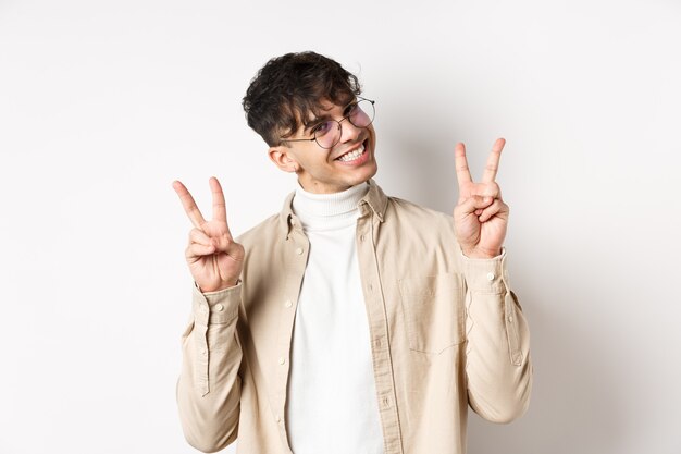 Echte Menschen. Hübscher Hipster-Typ mit Brille, der Friedensgesten zeigt und süß lächelt und auf weißem Hintergrund steht.