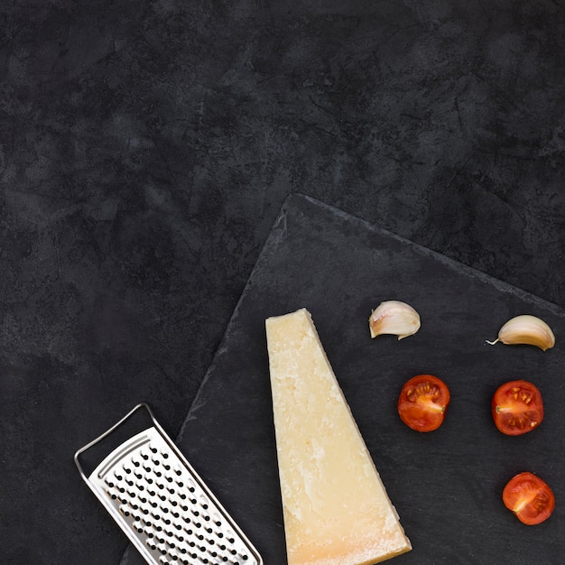 Kostenloses Foto edelstahlreibe mit käseblock; knoblauchzehen und halbierte tomaten auf schieferfelsen über dem schwarzen hintergrund