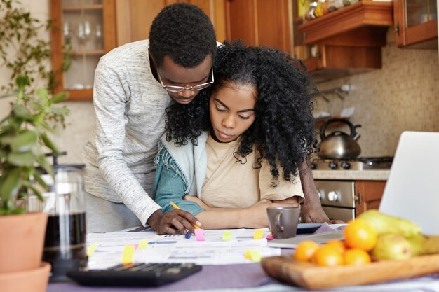 Ehemann in Gläsern hilft seiner schönen Frau beim Papierkram, steht neben ihr und erklärt etwas auf Papieren. Junge afrikanische Familie, die Finanzen zusammen verwaltet und am Küchentisch sitzt