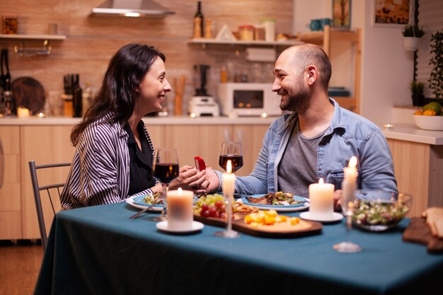 Ehemann schlägt seiner Frau vor, ihn während eines romantischen Abendessens in der Küche zu heiraten. Mann macht seiner Freundin beim romantischen Abendessen in der Küche einen Vorschlag. Glückliche kaukasische Frau, die sprachlos lächelt