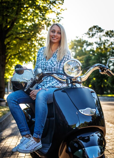 Ehrfürchtige blonde Frau in Jeans und Hemd, die auf Motoroller im grünen Park sitzt.