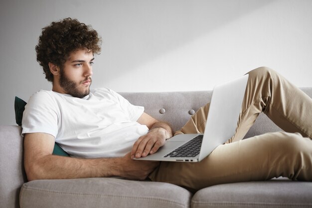 Ehrliche Aufnahme eines ernsthaften konzentrierten jungen unrasierten Mannes in einem weißen T-Shirt, das auf einem generischen Laptop im Internet surft, einen Film ansieht oder einen Online-Nachrichtenartikel liest, kostenloses WLAN nutzt und zu Hause auf der Couch sitzt