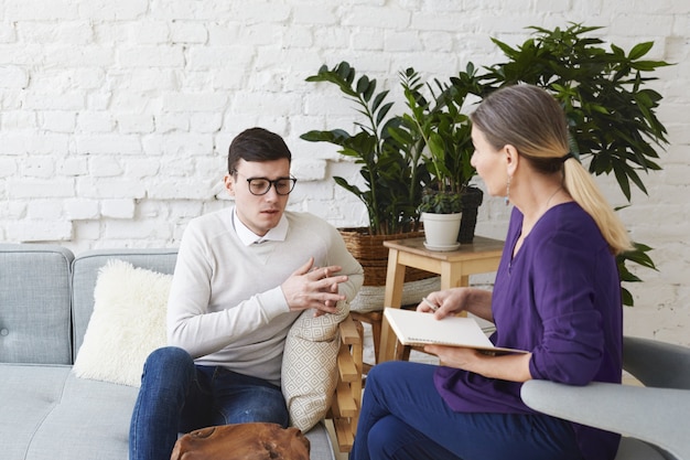 Ehrliche Aufnahme eines jungen Mannes in einer Brille, der über seine Probleme während einer psychologischen Therapiesitzung spricht, während er auf einer Kutsche sitzt, während eine reife Psychologin mit einem Heft ihm zuhört und sich Notizen macht