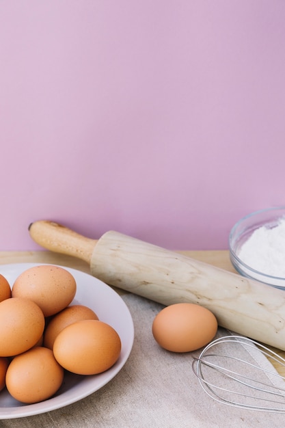 Eier auf Teller; Nudelholz; Schneebesen und Mehl auf dem Schreibtisch gegen rosa Hintergrund