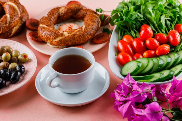 Eier mit wurst in einem teller mit einer tasse tee, türkischem bagel, oliven, salat, blumen auf rosa oberfläche