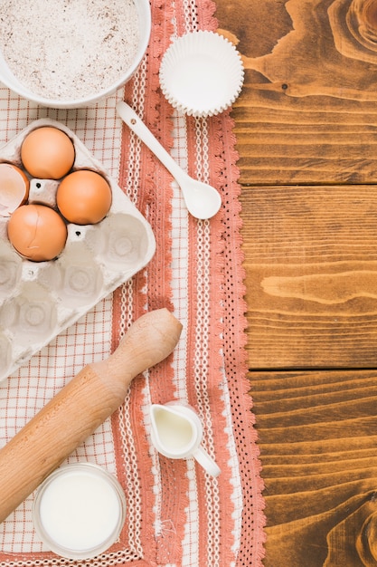 Kostenloses Foto eierkarton; mehlschüssel; milchglas; nudelholz; muffinform und löffel auf designertuch über strukturierter oberfläche