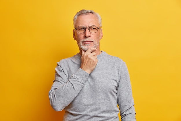 Ein älterer, durchsichtiger Mann hält das Kinn und schaut nachdenklich zur Seite. Die Planung trägt eine Brille und einen lässigen grauen Pullover, der über einer leuchtend gelben Wand isoliert ist