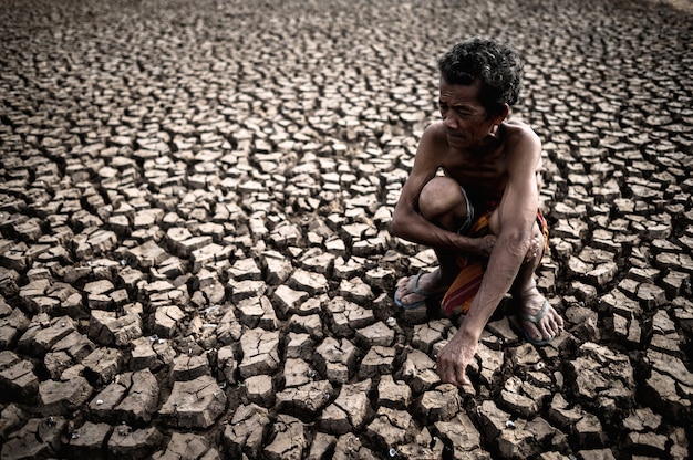 Ein älterer Mann, der seine Knie umarmend sitzt, verbog sich am unfruchtbaren Boden, globale Erwärmung