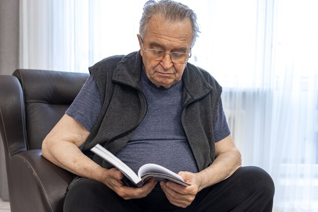 Ein älterer Mann mit Brille liest ein Buch, während er auf einem Sessel sitzt.