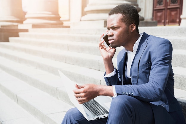 Ein afrikanischer junger Mann, der auf dem Treppenhaus hält den Laptop spricht auf Mobile sitzt