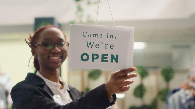 Ein afroamerikanischer Angestellter öffnet morgens ein Bekleidungsgeschäft und wartet auf Kunden. Ein BIPOC-Einzelhändler dreht ein geschlossenes Schild an der Tür einer Mode-Boutique im Einkaufszentrum.