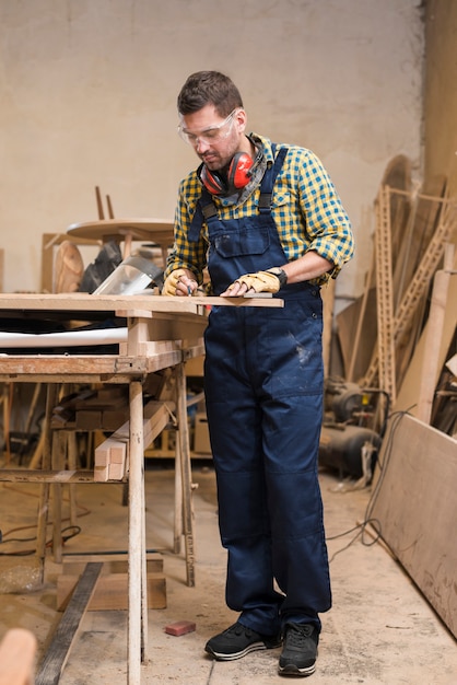 Ein aktiver männlicher Tischler, der die hölzerne Planke in der Werkstatt misst