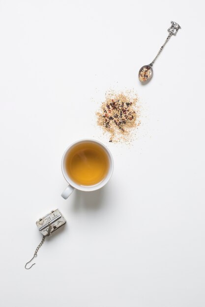 Ein altmodisches Teesieb; Löffel mit Kräutern und Tee in der Tasse auf weißem Hintergrund