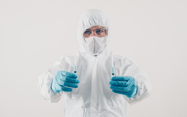 Ein Arzt hält 2 Spritzen mit Medizin in medizinischen Handschuhen und Schutzanzug in hellem Hintergrund.
