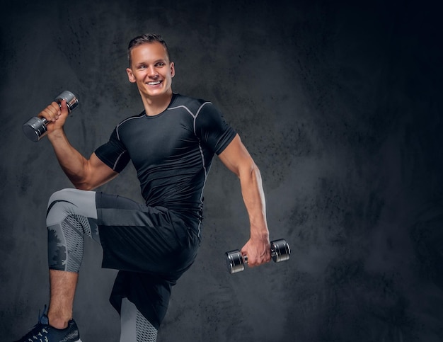 Ein athletischer Fitness-Mann in Sportbekleidung hält eine Reihe von Hanteln auf grauem Hintergrund.