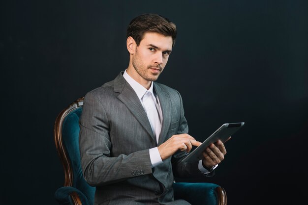 Ein attraktiver junger Geschäftsmann, der im Lehnsessel unter Verwendung der digitalen Tablette sitzt