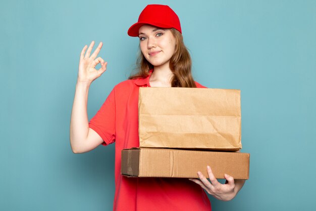 Ein attraktiver Kurier der Vorderansicht weiblicher in der roten Kappe des roten Poloshirts, die braune Pakete hält, die auf dem blauen Hintergrundlebensmittelservicejob lächeln