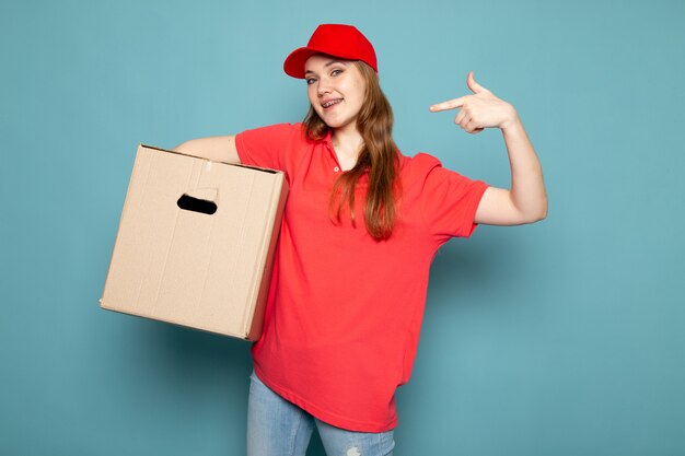 Ein attraktiver Kurier der Vorderansicht weiblicher in der roten Kappe des roten Poloshirts und in der Jeans, die Box hält, die lächelnd auf dem blauen Hintergrundlebensmittelservicejob aufwirft