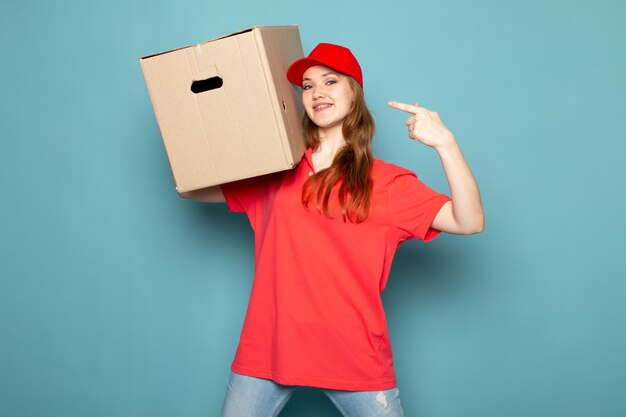 Ein attraktiver Kurier der Vorderansicht weiblicher in der roten Kappe des roten Poloshirts und in der Jeans, die Box hält, die lächelnd auf dem blauen Hintergrundlebensmittelservicejob aufwirft