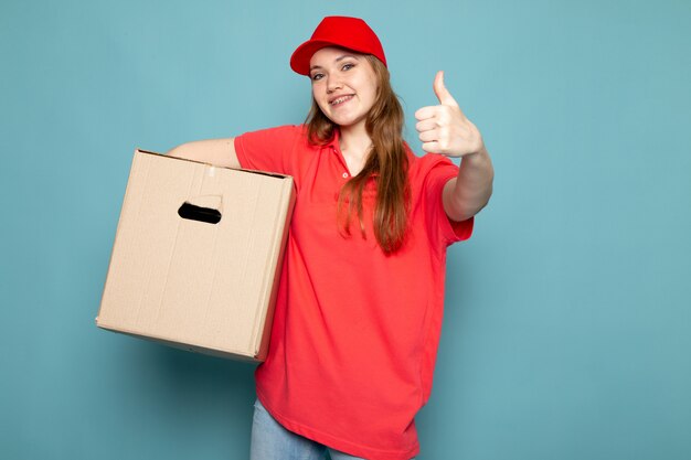 Ein attraktiver Kurier der Vorderansicht weiblicher in der roten Kappe des roten Poloshirts und in der Jeans, die Box hält, die lächelnd auf dem blauen Hintergrundlebensmittelservicejob aufwirft