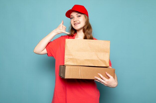 Ein attraktiver Kurier der weiblichen Vorderansicht in der roten Kappe des roten Poloshirts, die braunes Paket hält, unter Verwendung ihres imaginären Telefons, das auf dem blauen Hintergrundlebensmittelservicejob lächelt