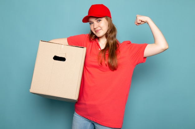 Ein attraktiver Kurier der weiblichen Vorderansicht in der roten Kappe des roten Poloshirts und in den Jeans, die Haltebox biegen, der auf dem blauen Hintergrundlebensmittelservicejob aufwirft