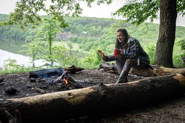 Ein attraktives Mädchen mit einer Tasse in der Hand sitzt auf einem Baumstamm und wärmt sich in der Nähe eines Feuers im Wald.