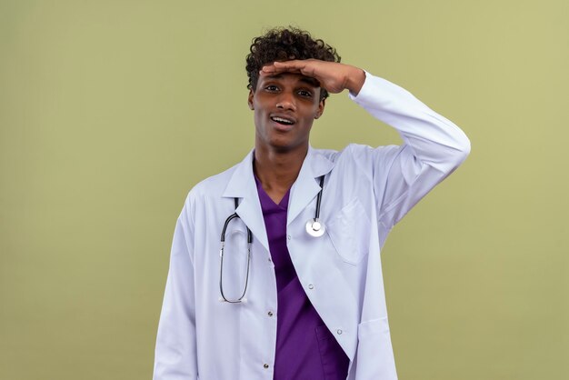 Ein aufgeregter junger gutaussehender dunkelhäutiger Mann mit lockigem Haar, der weißen Mantel mit Stethoskop trägt und sich auf eine Grünfläche freut