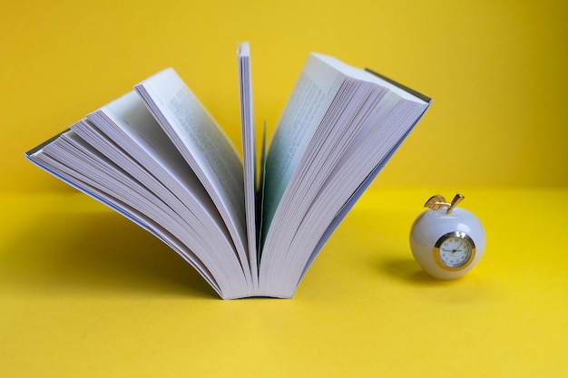 Ein aufgeschlagenes Buch auf gelbem Hintergrund mit einer Uhr in Form eines Apfels daneben