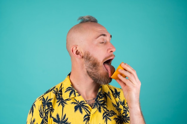 Ein bärtiger Mann in einem Hemd mit Palmenprint auf einer türkisfarbenen Wand leckt eine Orange