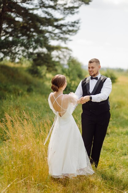 Ein bärtiger, stilvoller Bräutigam im Anzug und eine schöne blonde Braut in einem weißen Kleid mit einem Strauß in den Händen stehen und umarmen sich in der Natur im Kiefernwald.