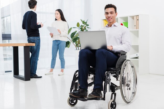 Ein behinderter lächelnder junger Geschäftsmann, der auf Rollstuhl mit Laptop vor Geschäftskollegen sitzt