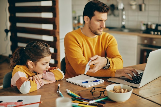 Ein beschäftigter Vater, der am Laptop arbeitet, während er seiner Tochter zu Hause etwas auf dem Smartphone zeigt