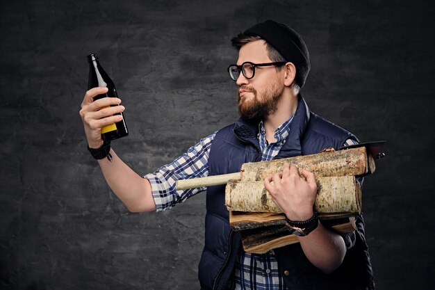 Ein betrunkener bärtiger Mann in einem Fleecehemd hält Feuerhölzer in seinen Armen und trinkt Craft-Flaschenbier.