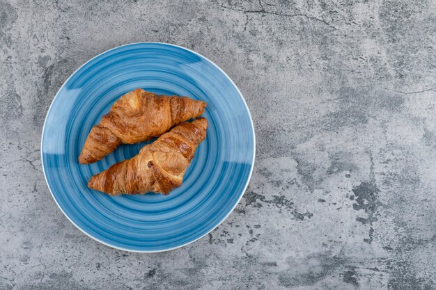 Ein blauer Teller mit zwei einfachen frischen Croissants auf einem Steintisch.