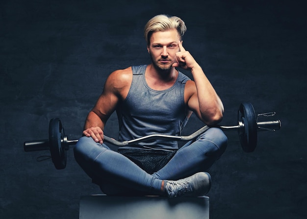 Ein blonder, athletischer Mann in grauer Sportkleidung sitzt auf einer weißen Holzkiste und hält eine Barbe.