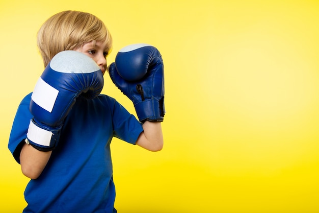 Ein blonder Junge der Vorderansicht, der Boxen in blauen Boxhandschuhen auf der gelben Wand aufwirft