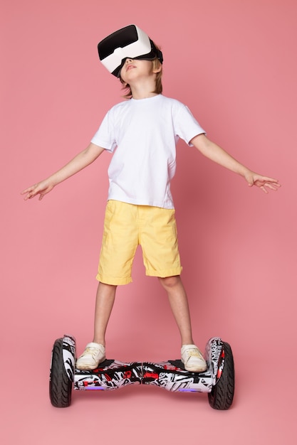 Kostenloses Foto ein blonder junge der vorderansicht im weißen t-shirt, das vr auf segway auf dem rosa raum spielt