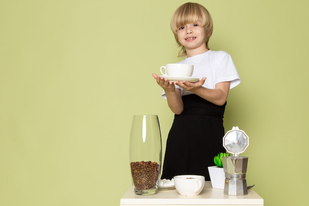 Ein blonder kinderjunge der vorderansicht im weißen t-shirt, das kaffeegetränk auf dem steinfarbenen raum vorbereitet